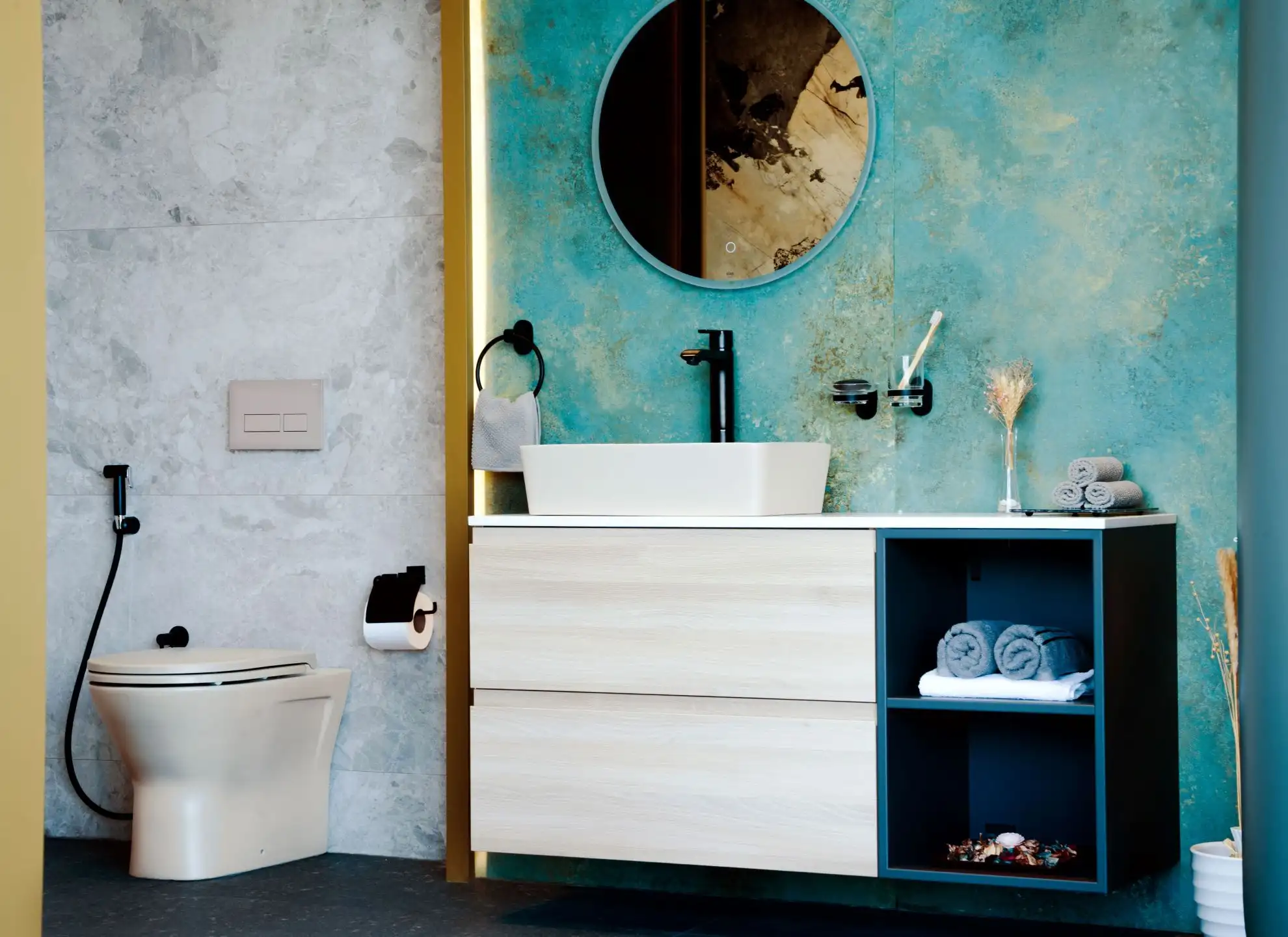 Modern white toilet and floating vanity against a split textured wall of blue-green and grey marble tiles with a circular mirror.